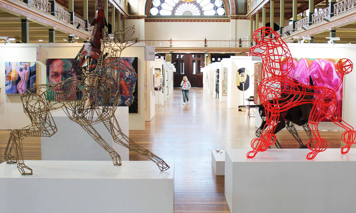 Photograph showcasing intricate dog sculptures by Cezary Stulgis, crafted from wood and metal, displayed prominently at the inaugural Melbourne Affordable Art Fair. The sculptures are juxtaposed with diverse artwork on the surrounding walls in a spacious, well-lit gallery with architectural details.