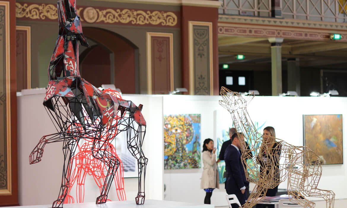 Photograph featuring a detailed sculpture by Cezary Stulgis, composed of metal and vibrantly colored fragments. The sculpture resembles a dog and stands in the foreground of a spacious art gallery adorned with intricate architectural designs. In the background, attendees converse near a variety of displayed artworks.