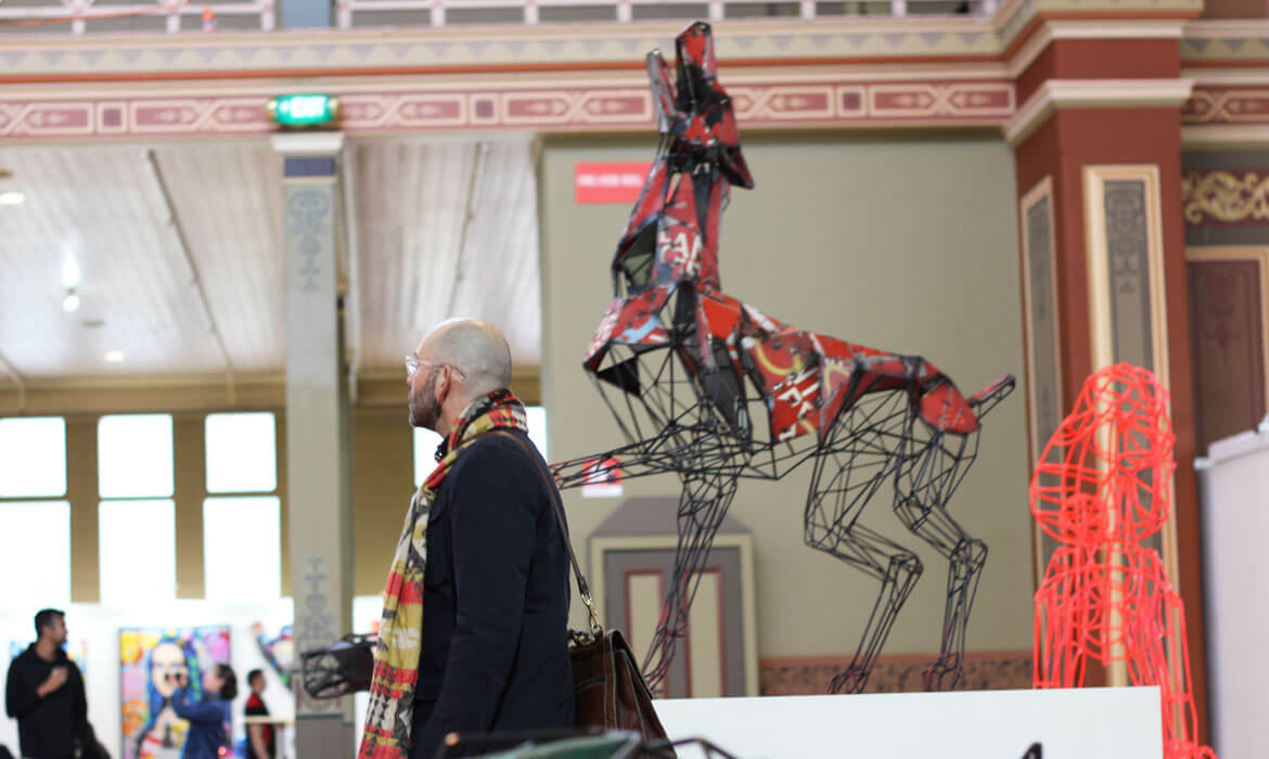 Photograph of an art gallery interior where a man with a colorful scarf and glasses observes a distinctive metal sculpture by Cezary Stulgis, resembling a dog made from vibrant fragments and wireframes. The expansive gallery has classical architectural details, and other artworks and visitors can be seen in the background.