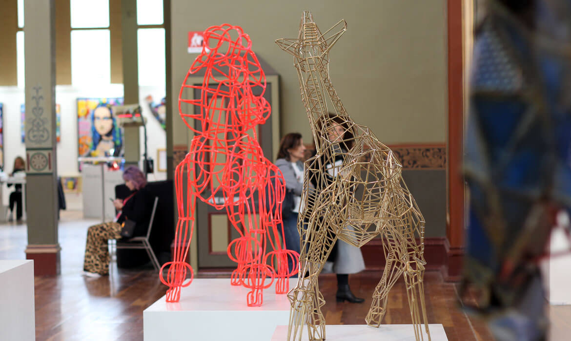 Photograph of an art exhibition hall showcasing wireframe sculptures by Cezary Stulgis: a vibrant red humanoid figure and a gold-colored giraffe, both intricately designed. Visitors explore the space, with a woman seated sketching nearby, and various other art pieces are visible in the background, including a portrait painting and ornate architectural columns.