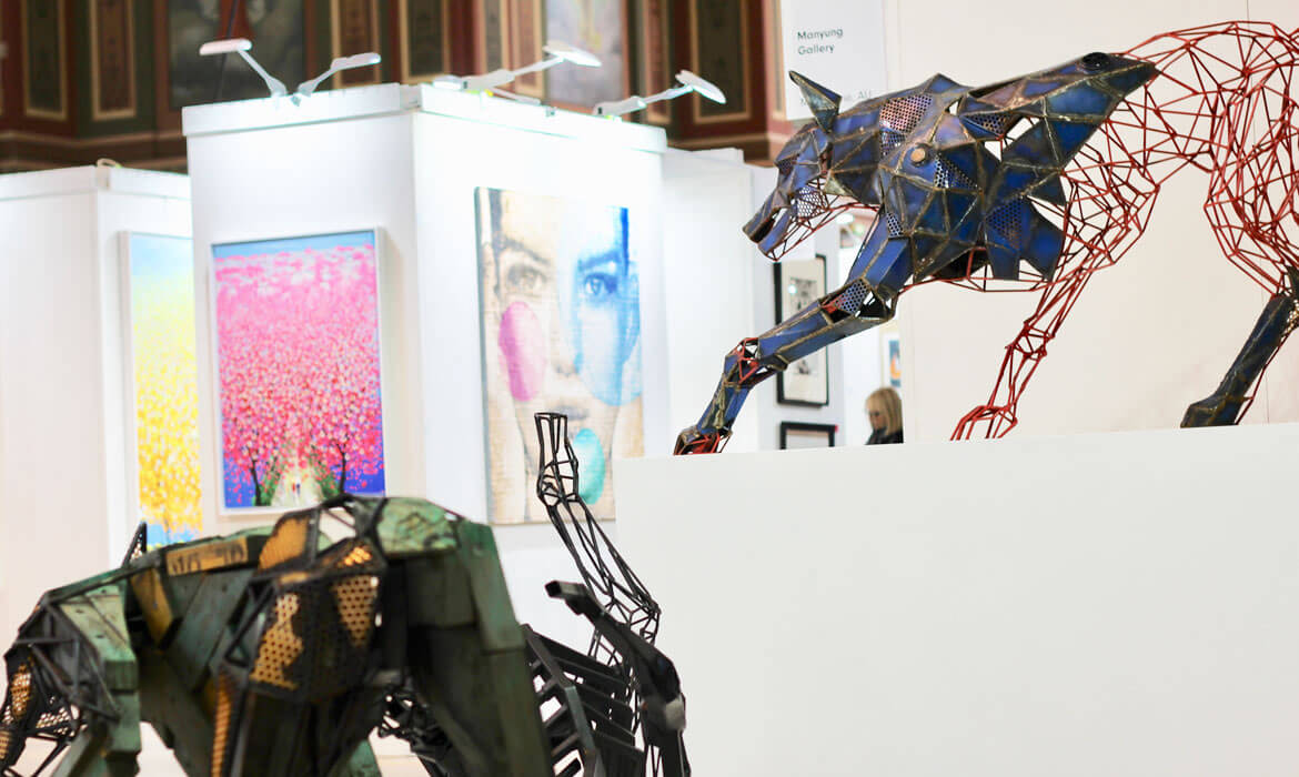 Photograph of an art exhibition interior featuring a dynamic wireframe sculpture by Cezary Stulgis: a large blue and red canine form, its head turned and jaws open. In the foreground, there's a partial view of another sculpture resembling a horse. The walls showcase colorful paintings, including a vibrant tree blossoming in pink and a fragmented portrait. A gallery banner and a visitor are also visible in the background.