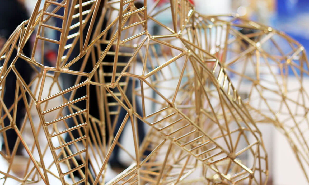 Close-up photograph of a golden wireframe sculpture by Cezary Stulgis, showcasing intricate geometric patterns and intersections. The gleaming metallic rods form a detailed mesh, with blurred hints of the exhibition space in the background.