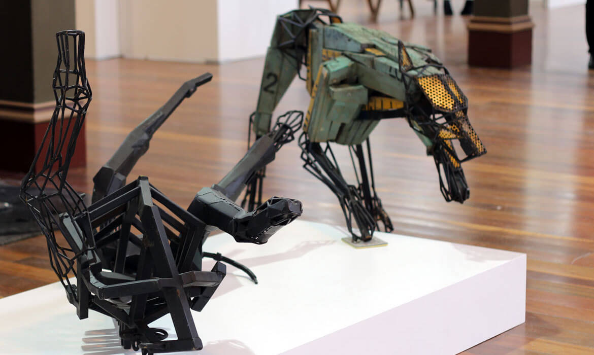 Photograph of two distinct sculptures by Cezary Stulgis set in an indoor exhibition space. The foreground features a complex black wireframe structure with geometric interplay, reminiscent of shattered or deconstructed elements. In the background, a meticulously crafted mechanical creature resembling a dog or wolf, adorned with green plates, intricate wire mesh, and yellow accents, stands poised on a white pedestal against a polished wooden floor.