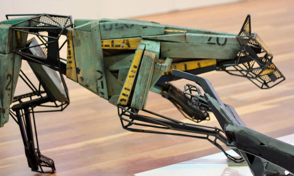 Close-up view of a Cezary Stulgis sculpture, depicting a mechanical creature's detailed body. Crafted with aged green metal panels, featuring markings like '20' and yellow caution stripes, the sculpture seamlessly integrates wireframe elements. Parts of its skeletal structure and muzzle, made from black wire mesh, contrast against the solid panels. The artwork stands against a backdrop of a light-colored wall and polished wooden floor.