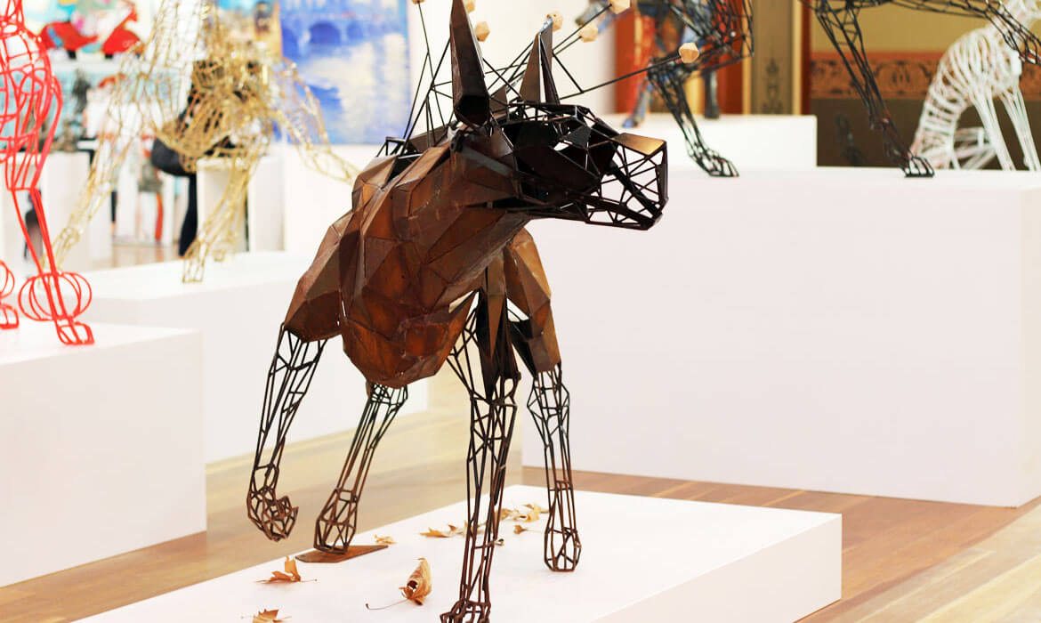 Foreground showcases a Cezary Stulgis sculpture resembling an animal with elongated legs and a protruding snout. The sculpture is constructed using a combination of rusty brown, faceted metal panels for its body and black wire mesh detailing for its limbs, muzzle, and ears. Autumn leaves are scattered at its feet. In the blurred background, various other abstract sculptures in red, gold, and white hues can be seen displayed in a well-lit gallery space with a hint of wall art.