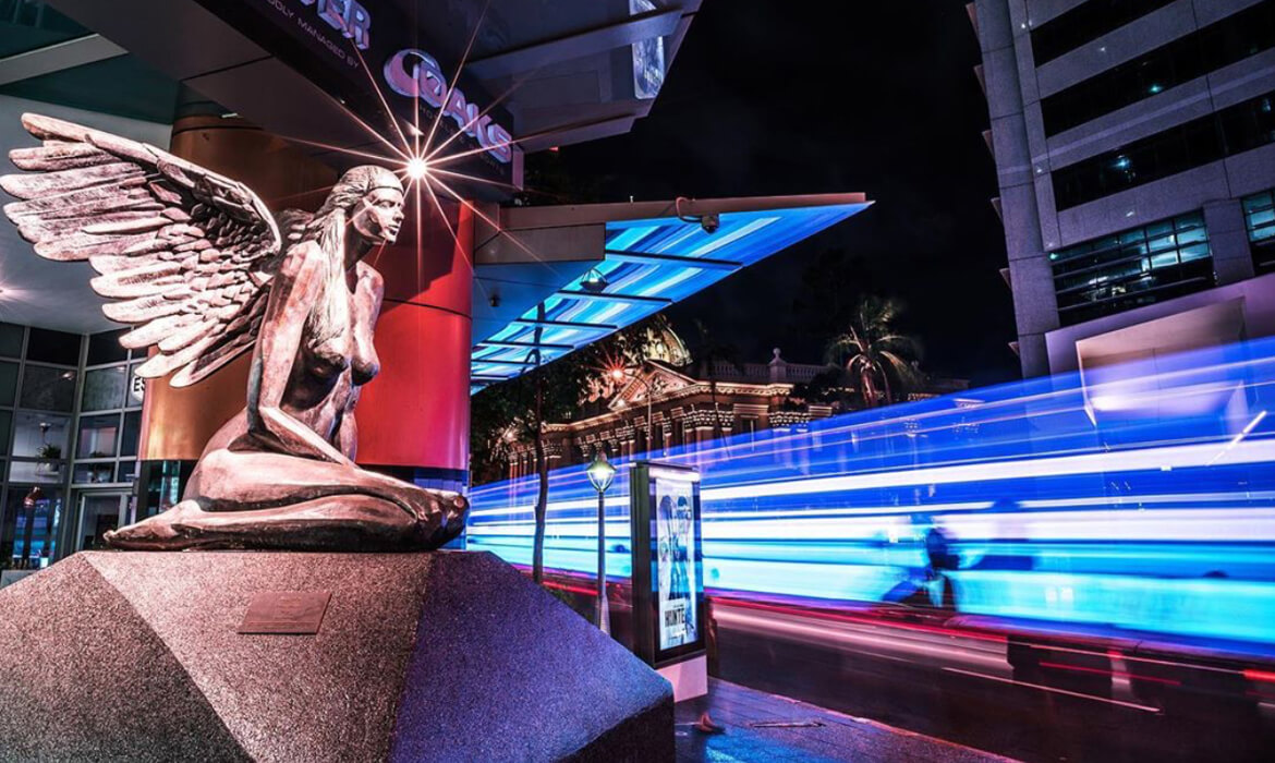 A stunning bronze sculpture titled 'Aurora' depicting a kneeling winged woman, illuminated against the backdrop of a bustling urban setting at night. The figure's detailed wings stretch outward as she seems to be in deep thought or prayer. Behind her, the vibrant blue and white streaks of traffic light trails contrast with the red hue of the nearby building's facade. Further in the distance, a modern building stands tall with lit windows, and to the left, a tropical palm tree and part of a historic building can be seen