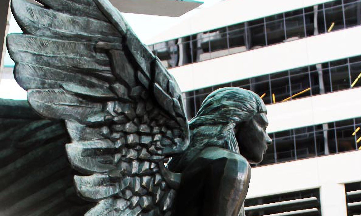 A close-up view of a bronze sculpture titled 'Aurora', showcasing a detailed wing with intricate feather patterns. The wing extends from a female figure's back, with her head turned to the side, revealing her delicate facial features and flowing hair. In the background, the monochrome facade of a modern building is seen, with rectangular windows reflecting the ambient light.