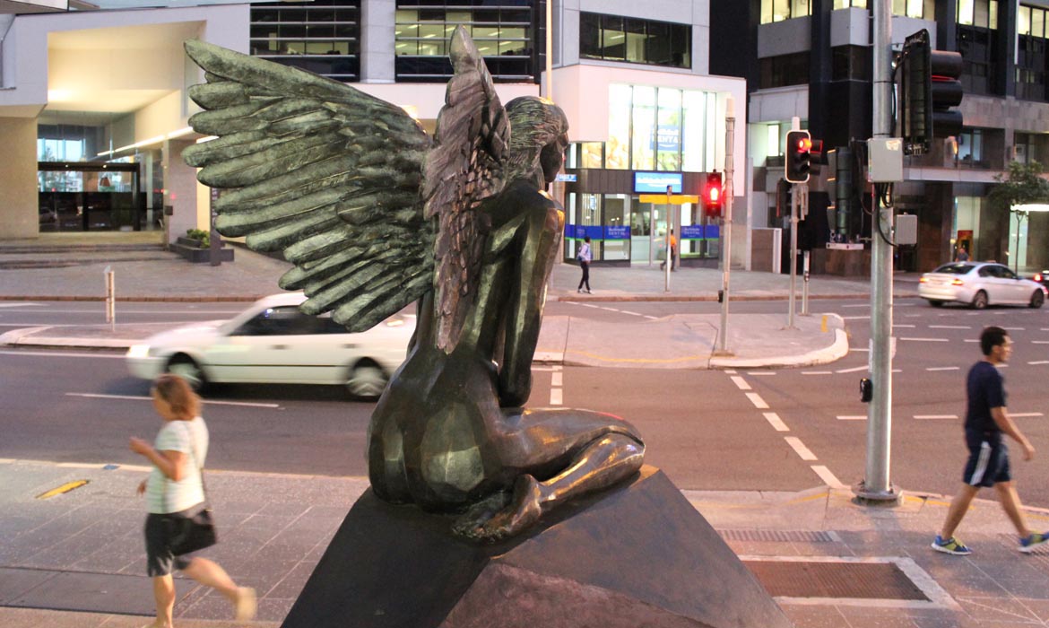 A bronze sculpture titled 'Aurora' is prominently displayed at an urban intersection. The artwork features a female figure with large, detailed wings extended. She appears to be seated or kneeling on a pedestal. In the background, pedestrians cross the street, cars pass by, and a modern building with lit windows and a bank facade is visible. The scene captures a blend of art and daily city life.