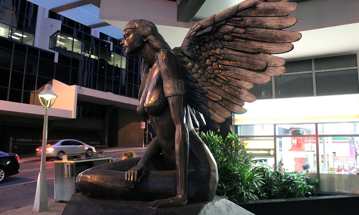 A close-up view of a bronze sculpture named 'Aurora' set against an urban backdrop. The sculpture showcases a graceful female figure with intricately detailed wings spread behind her. She is poised with one knee raised, displaying a sense of movement. Behind Aurora, cars blur in motion, a retro-styled streetlamp stands tall, and a modern building with large windows and a brightly lit store is visible, illustrating the melding of art and city life.