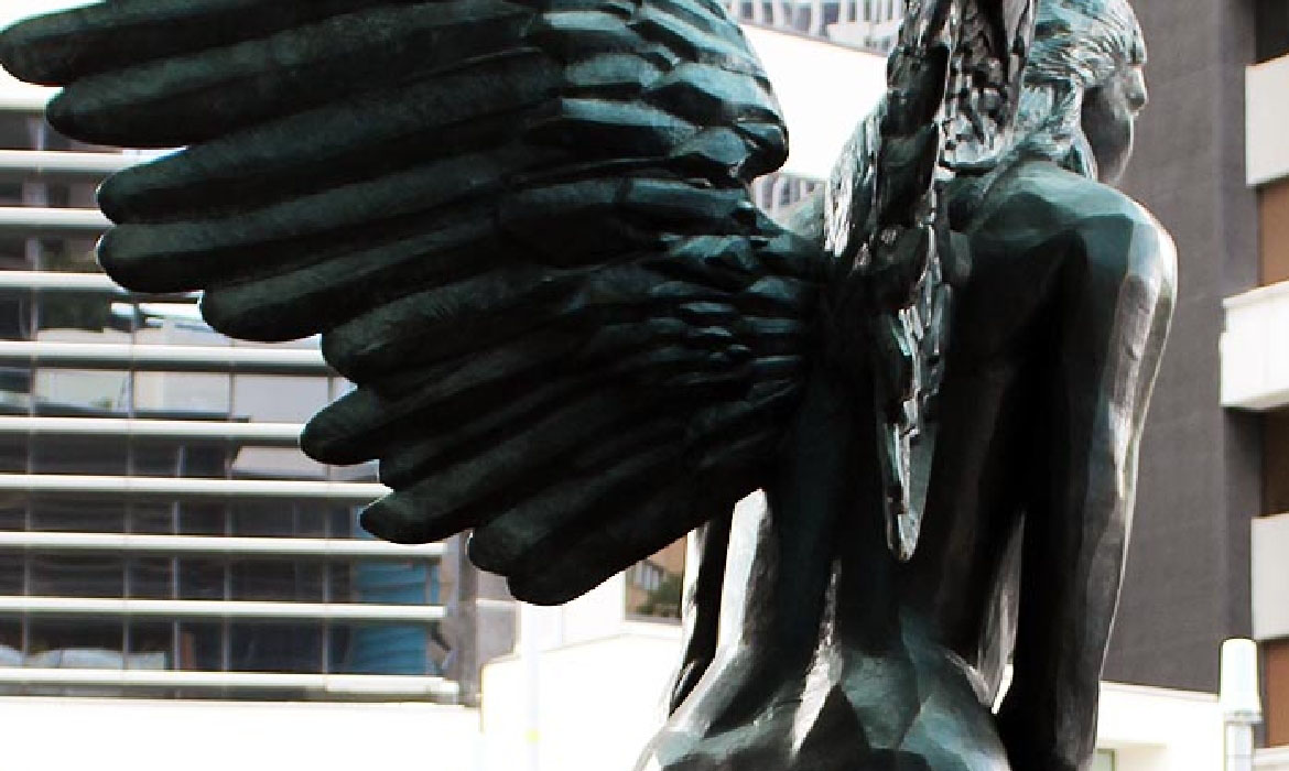 A zoomed-in detail of the 'Aurora' sculpture, emphasizing the textured wings and sleek form of the female figure. The bronzed, feathered wings radiate an aura of strength and beauty. In the background, the reflective windows of a modern city building offer a stark contrast to the organic form of the sculpture, capturing the intersection of art and urban architecture.