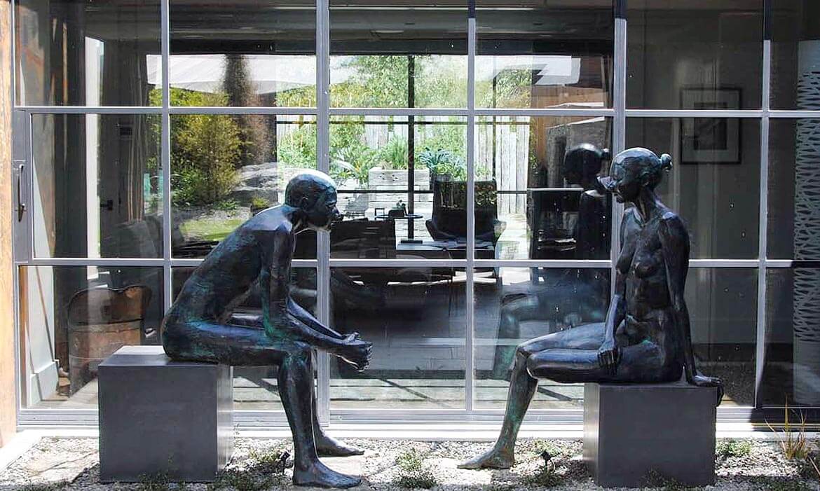 A pair of bronze sculptures titled 'The Conversation' positioned in front of large glass windows. The male figure sits on the left, with his hands clasped, gazing forward. The female figure on the right sits with one leg folded, turning slightly towards the male figure. Behind them, the glass windows reveal an interior space with modern furnishings and an outdoor garden reflecting the serene environment in which they're placed.