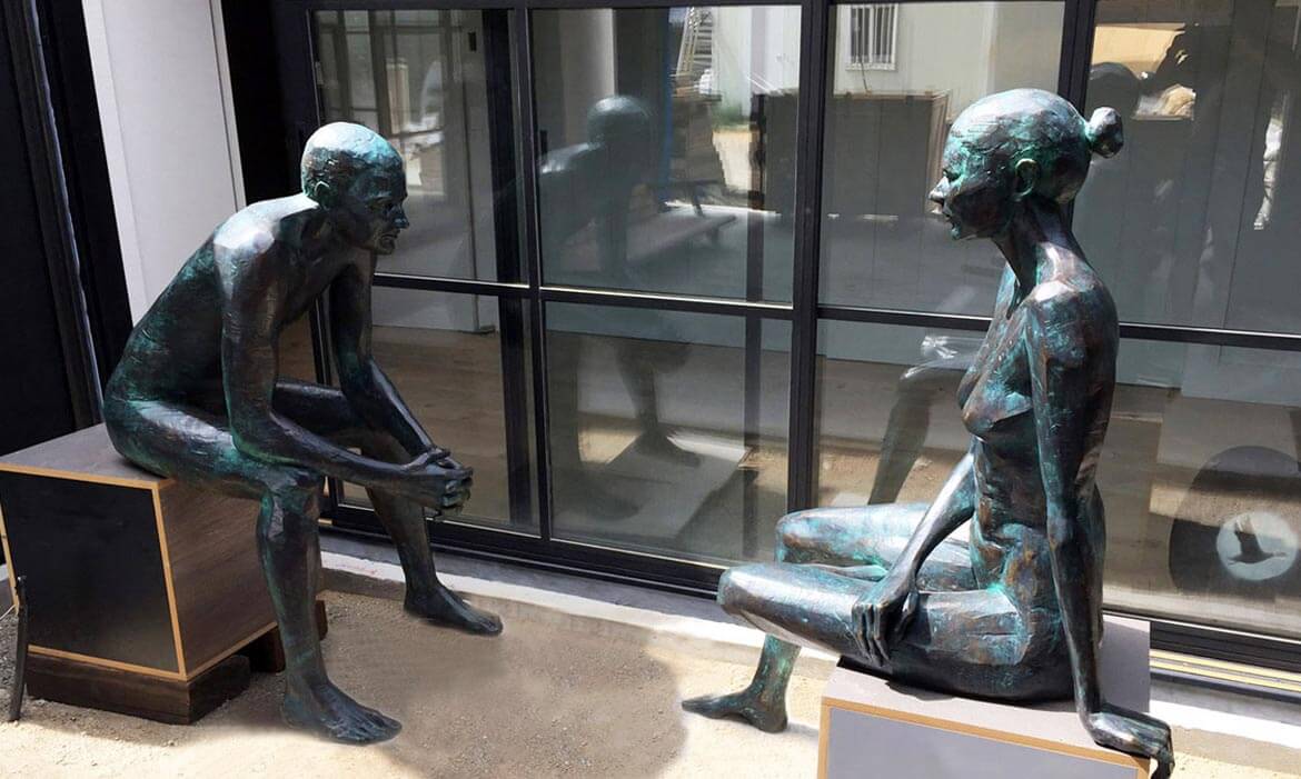 Two bronze sculptures titled 'The Conversation' placed near large sliding glass doors. The male sculpture on the left is seated on a wooden block, hands clasped in deep thought, while the female sculpture on the right sits gracefully on another block with one leg folded and her gaze directed forward. Their shiny and patinated surfaces reflect the surroundings, and through the glass doors, a glimpse of an interior space can be seen.