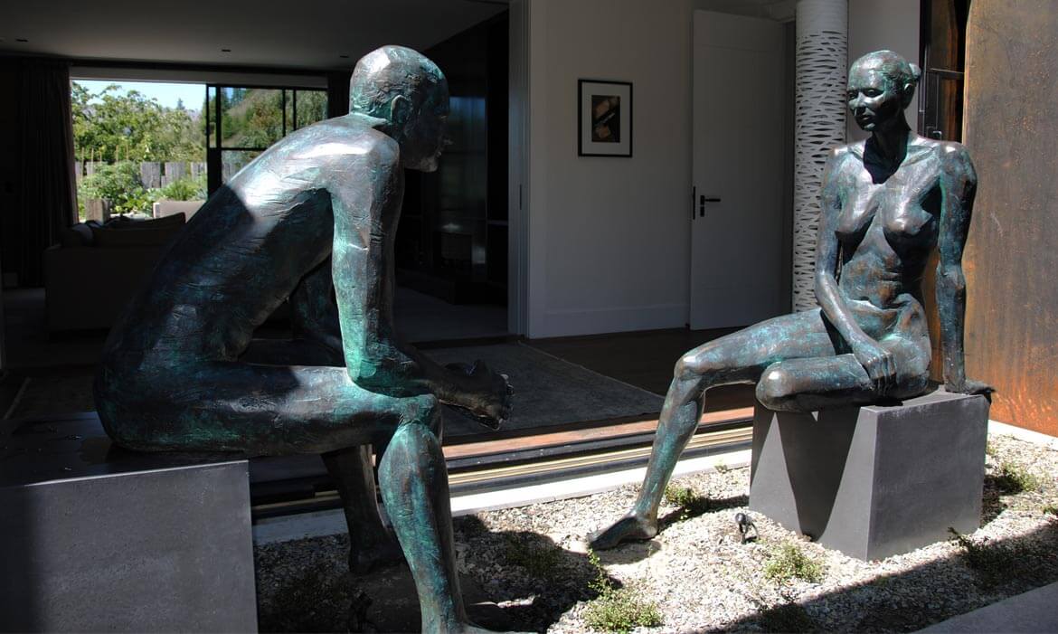 Two detailed bronze sculptures titled 'The Conversation' positioned outside a modern home with large glass doors. The male sculpture on the left sits contemplatively on a concrete base, with one knee raised and hands clasped, facing the female sculpture on the right. She rests gracefully on a similar concrete base, one leg folded beneath her. Behind them, the interior of the home is visible, with contemporary furniture and artwork adorning the walls.