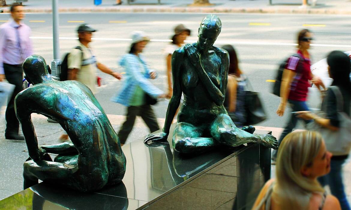 Bronze sculptures titled 'Dialogue' depict two human figures in contemplative poses on a city street, with blurred pedestrians walking past in the background.