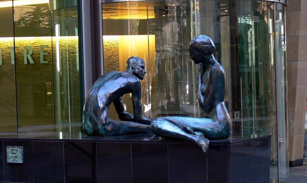 Close-up view of the bronze sculptures titled 'Dialogue', featuring two human figures seated face to face in a deep conversation, set against a backdrop of a modern building with reflective glass doors and walls.