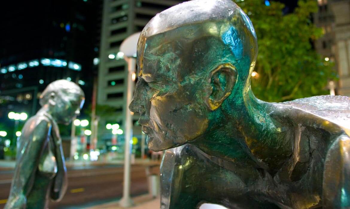 Close-up of the bronze sculpture titled 'Dialogue', showcasing the intricate details of a male figure's face, with another blurred figure in the background. The scene is illuminated by ambient city lights, with urban buildings and trees visible in the distance.