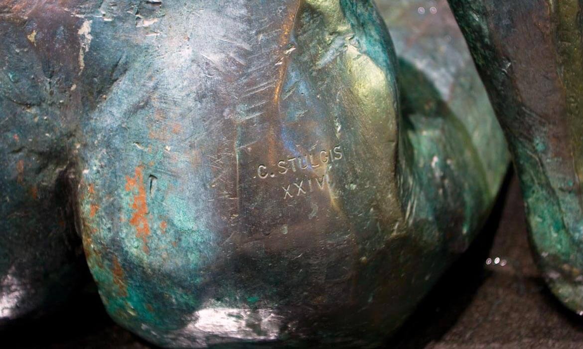 Close-up view of the patinated surface of the 'Dialogue' sculpture, focusing on an engraved signature reading 'C. STULGIS' followed by Roman numerals 'XXIV'. The texture reveals hints of rust and wear, hinting at the passage of time.