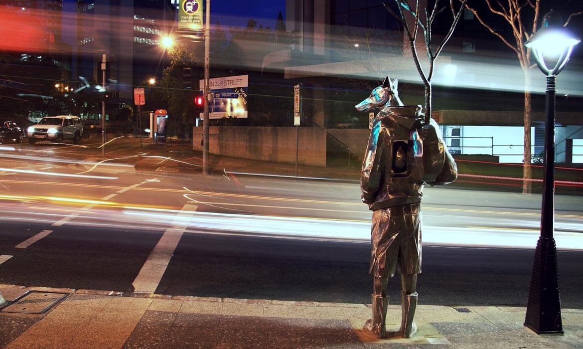 A metallic sculpture, 'The Guardian',  of a humanoid figure with fox-like features stands by a city street at nighttime. The sculpture is adorned with a backpack and holds onto a pole. Streaks of light from passing traffic create a dynamic contrast against the stillness of the figure, while illuminated buildings and a vintage street lamp add depth to the urban backdrop.