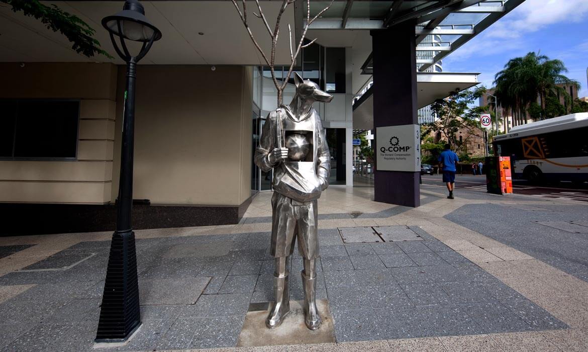 A metallic sculpture, 'The Guardian',  of a humanoid figure with fox-like facial features stands on a city sidewalk. The sculpture holds a thin, bare tree and has a sphere embedded in its torso. Adjacent to it stands a street lamp, with a building's facade in the background. In the distance, a pedestrian walks by, and a bus is visible near a sign that reads 'Q-COMP'.