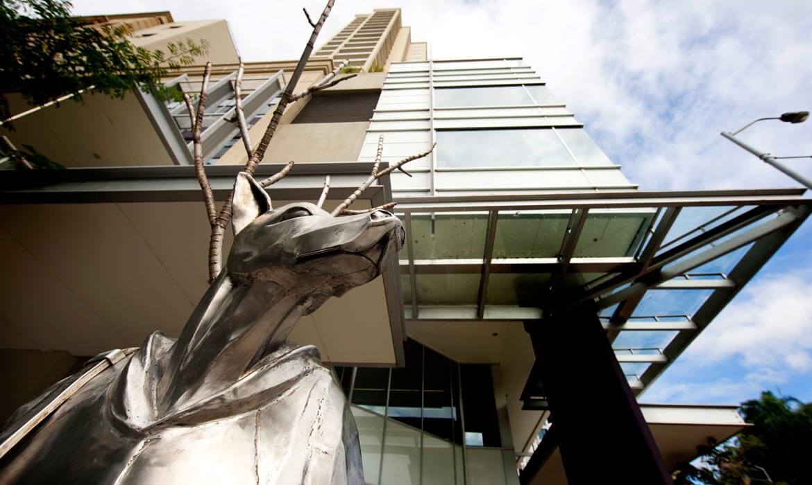 Close-up view of a metallic dog sculpture, 'The Guardian', with bare tree branches, set against modern urban buildings and a blue sky with white clouds.