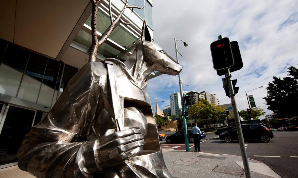 Metallic dog sculpture named 'The Guardian' positioned near a street with a pedestrian crosswalk signal, buildings in the background, and a pedestrian waiting to cross the road.