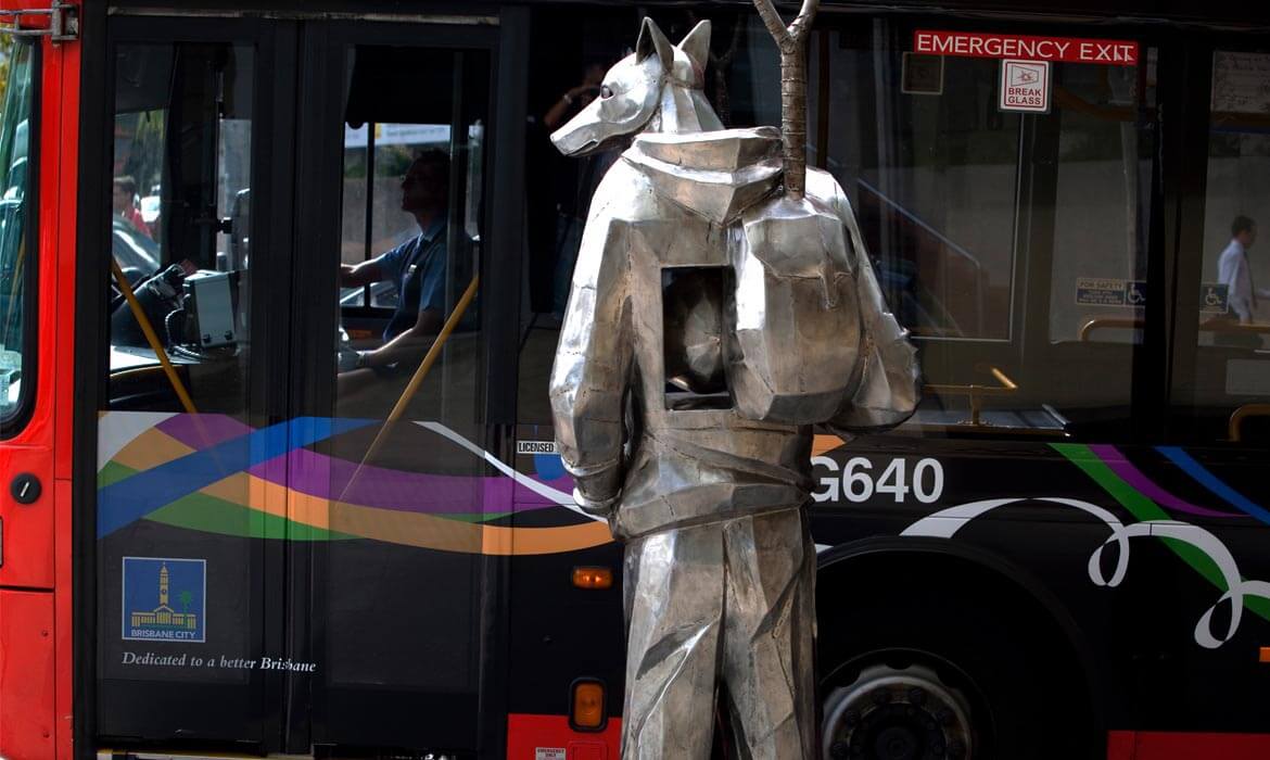 Metallic dog sculpture named 'The Guardian' stands beside a colorful city bus with the text 'Dedicated to a better Brisbane' and a bus driver visible through the door window.