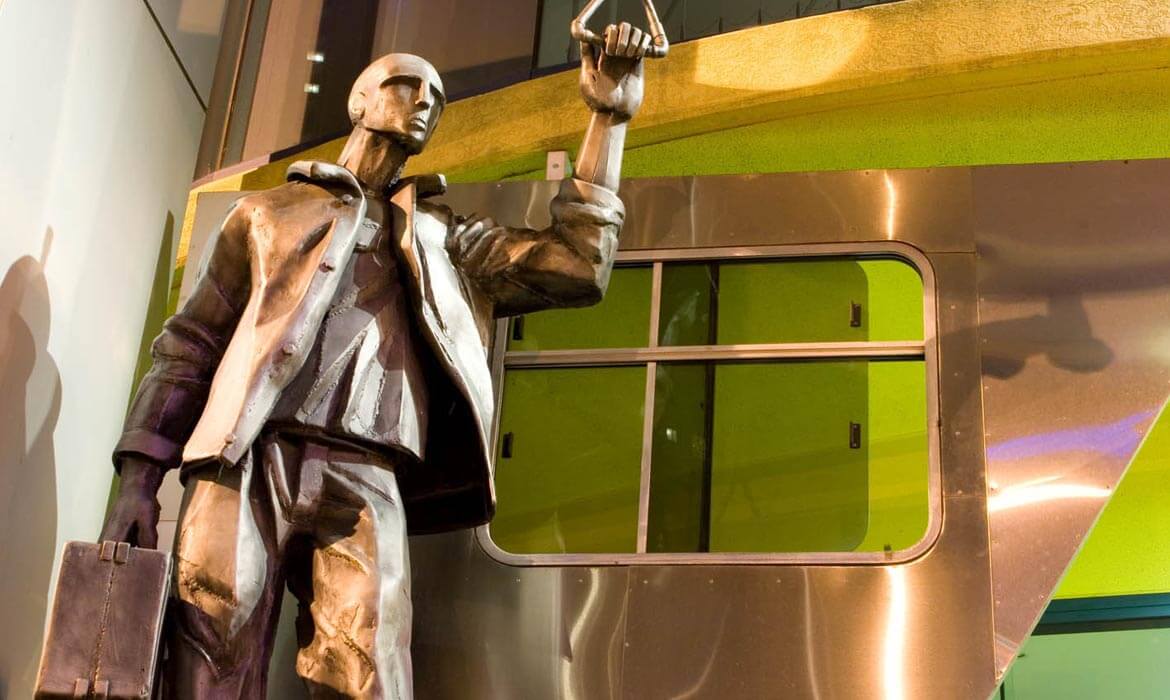 Sculpture of a man in a coat holding onto a transportation handle, standing next to a metallic structure with green windows.