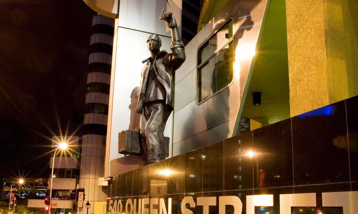 Nighttime view of a large bronze statue of a man holding onto a transportation handle, adjacent to a building labeled '540 QUEEN STREET', with bright streetlights illuminating the scene.