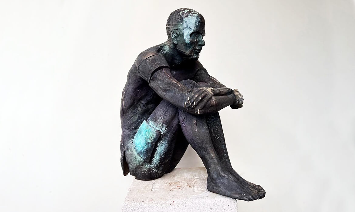 Art piece titled 'Replica001' by Cezary Stulgis. A cast bronze sculpture measuring 550mm x 470mm x 260mm, displayed in Melbourne, Australia. The sculpture depicts a dark, patinaed human figure with blue-green oxidation spots, sitting contemplatively with crossed legs, hands resting on the ankles. The intricately detailed figure exudes a sense of deep thought and introspection, set against a light neutral background.