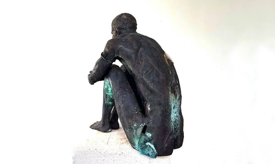 Art piece titled 'Replica001'. A cast bronze sculpture displaying the rear view of a human figure. The sculpture showcases a dark, patinaed surface with hints of blue-green oxidation, particularly on the lower half. The figure appears in a hunched, introspective posture, emphasizing the natural and intricate details of the work. Set against a light neutral background, the piece evokes a sense of contemplation and solitude.