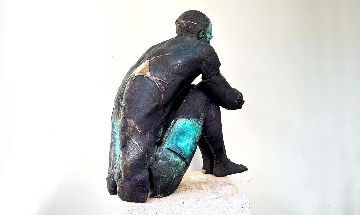 Art piece titled 'Replica001'. Bronze sculpture of a human figure seen from a side-angle, displaying intricate detailing and textures. The figure is captured in a reflective pose, sitting with its hands resting on the knees. Dark patina covers most of the surface with vibrant blue-green oxidation highlights on the legs and back. The sculpture is set against a pale, neutral background, enhancing the visual depth and contemplative nature of the piece