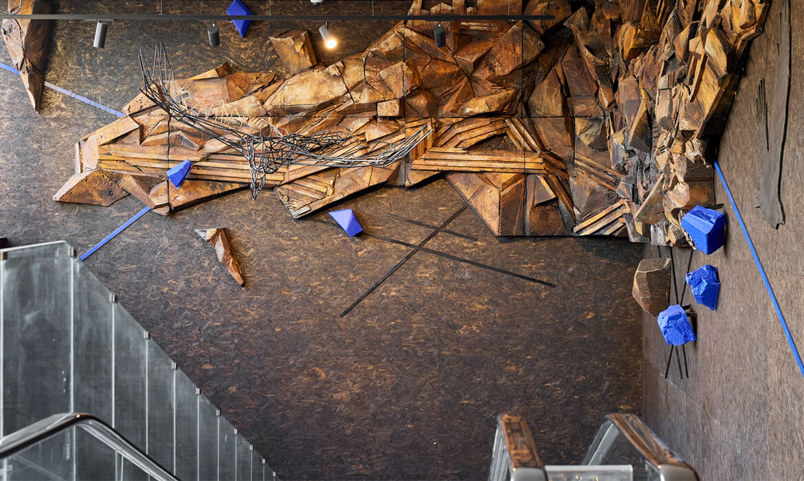 A detailed section of an art installation titled 'Terrain'. The artwork showcases multi-layered geometric formations, resembling rugged mountainous terrains, made from textured and painted styrofoam in deep browns and golds. There are vibrant blue accents scattered throughout. On the dark, patterned floor, there's a silhouette of a bird in mid-flight, providing a stark contrast. The overall aesthetic evokes a sense of dynamic topography and nature's intricate designs.