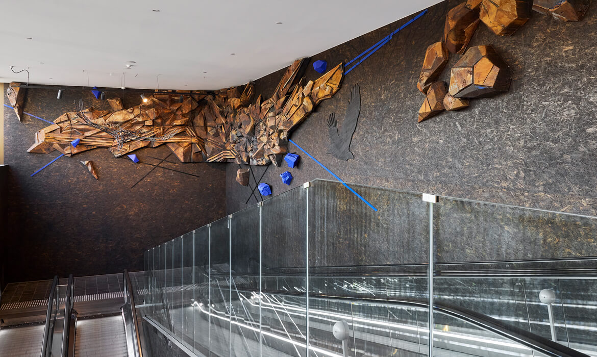 An overhead view of the 'Terrain' art installation. It features a multi-dimensional landscape made from intricately layered and painted styrofoam, resembling rugged mountainous terrains with rich brown and gold hues. Intermingled are abstract wire sculptures and striking blue geometric accents that stand out. The patterned dark floor complements the installation, and a sleek glass barrier is visible to the left, suggesting an observation or walking platform for viewers to engage with the artwork.