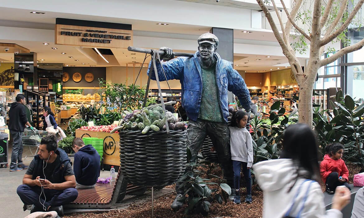 The 'Vegetable Hawker' art installation at High Point Westfield Plaza. A life-sized cast aluminium statue, painted with realistic details, depicts a hawker in a jacket and cap, holding a large basket brimming with sculpted vegetables. The artwork, measuring 220cm x 90cm x 200cm, stands prominently in the shopping plaza with shoppers, greenery, and the backdrop of a fruit and vegetable market. Crafted from aluminium, stainless steel, and paint, it was commissioned by GPT and completed in 2022.