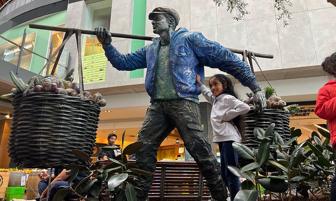 Close-up view of the 'Vegetable Hawker' sculpture at a shopping plaza. The life-sized statue showcases a man in a blue jacket and cap, striding forward with a large, intricately designed basket of sculpted vegetables balanced on a pole across his shoulders. A young girl stands beside him, looking up and reaching out to touch the artwork. Background features shoppers, a bench, green plants, and storefronts, emphasizing the interactive nature of this public art piece.