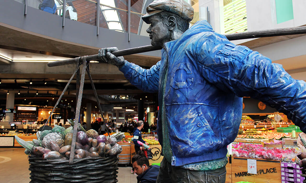 Side profile of the 'Vegetable Hawker' sculpture positioned inside a bustling marketplace. The sculpture depicts a man in a textured blue jacket and cap, carrying a basket filled with detailed sculpted vegetables suspended on a pole across his shoulder. In the backdrop, shoppers can be seen browsing fresh produce, with stalls displaying an array of colorful fruits and vegetables. Above, architectural elements like a mezzanine and skylights add depth to the scene.