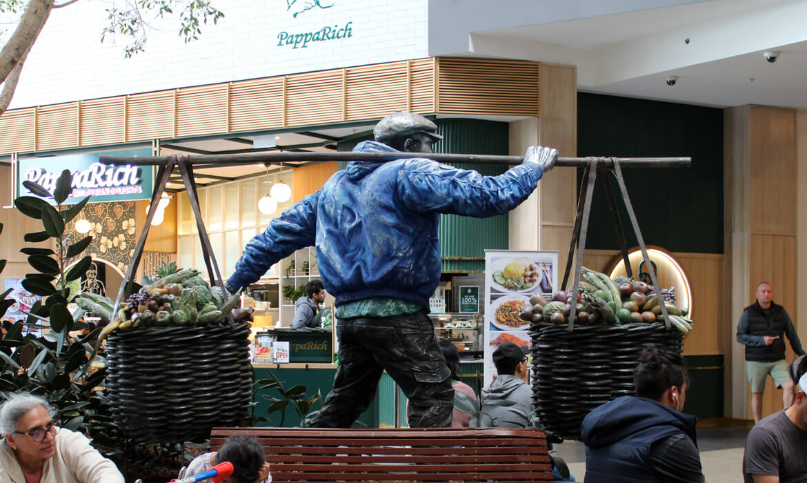 Rear view of the 'Vegetable Hawker' statue in a lively mall setting. The statue portrays a man in a detailed blue jacket, carrying two large baskets filled with intricately sculpted vegetables, balanced on a pole across his shoulders. In the background, the 'PappaRich' restaurant signage is prominently displayed, with patrons seated and dining nearby. Other shoppers can be observed walking or sitting, adding to the bustling atmosphere. Modern architectural designs, including wooden accents and pendant lights, contribute to the scene's ambiance.