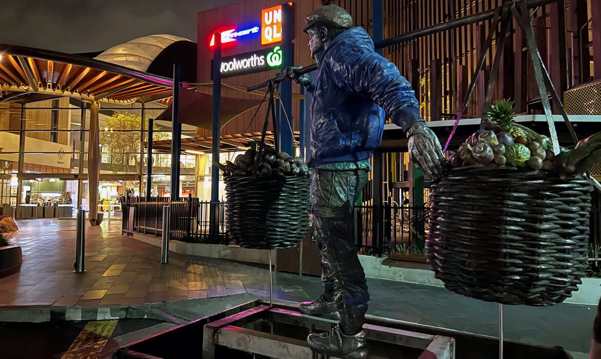 Nighttime view of the 'Vegetable Hawker' statue placed outside a shopping area. The lifelike statue, illuminated by surrounding lights, captures a man wearing a detailed blue jacket, holding two large baskets brimming with sculpted vegetables, balanced on a pole across his shoulders. Adjacent to the statue are neon signages of popular brands like 'UNIQLO' and 'Woolworths'. The scene is enhanced by architectural details such as modern steel canopies, decorative fencing, and a serene water feature below the statue's platform. Shops with warm interior lighting can be seen in the background.