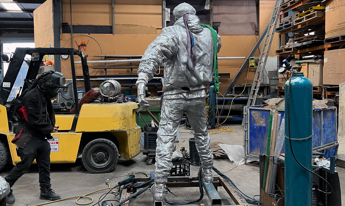 Inside a warehouse, a life-sized statue titled 'Vegetable Hawker' is in the process of being crafted. The statue depicts a man in detailed silver attire with a cloth draped over his shoulder, standing next to an industrial workspace. To the left, a worker wearing a black jacket and protective welding helmet is bent over a yellow forklift, inspecting machinery. Surrounding the scene are various tools, metal scraps, gas cylinders, and workshop equipment, with shelves stacked with boxes in the background. The ambiance is that of a busy and productive artistic workshop.